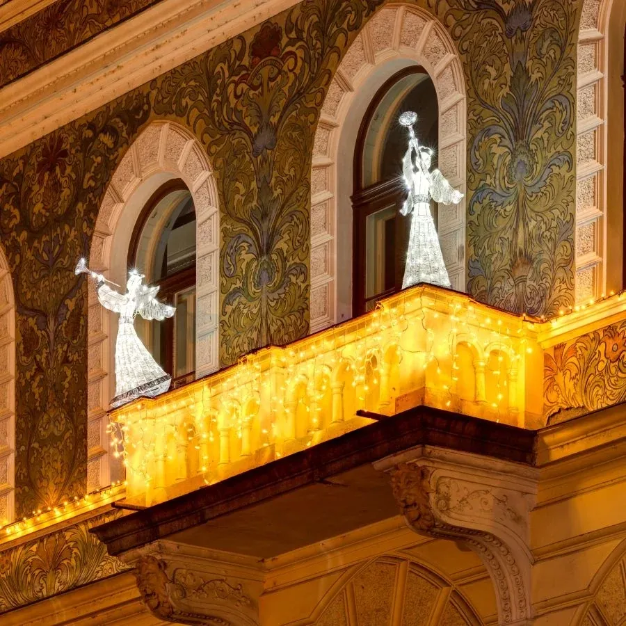 LED anjeli s trúbkou v ľadovo bielej na balkóne historickej fasády so svetelnými cencúľmi v teplej bielej